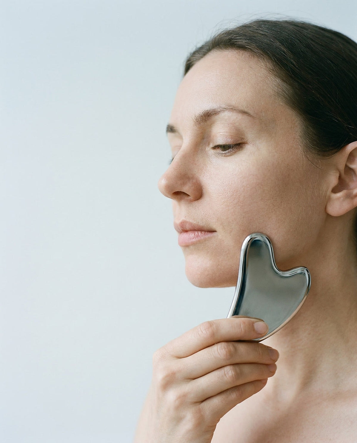 A woman gliding the polished stainless steel SENVÈ gua sha along her jaw against a soft grey background