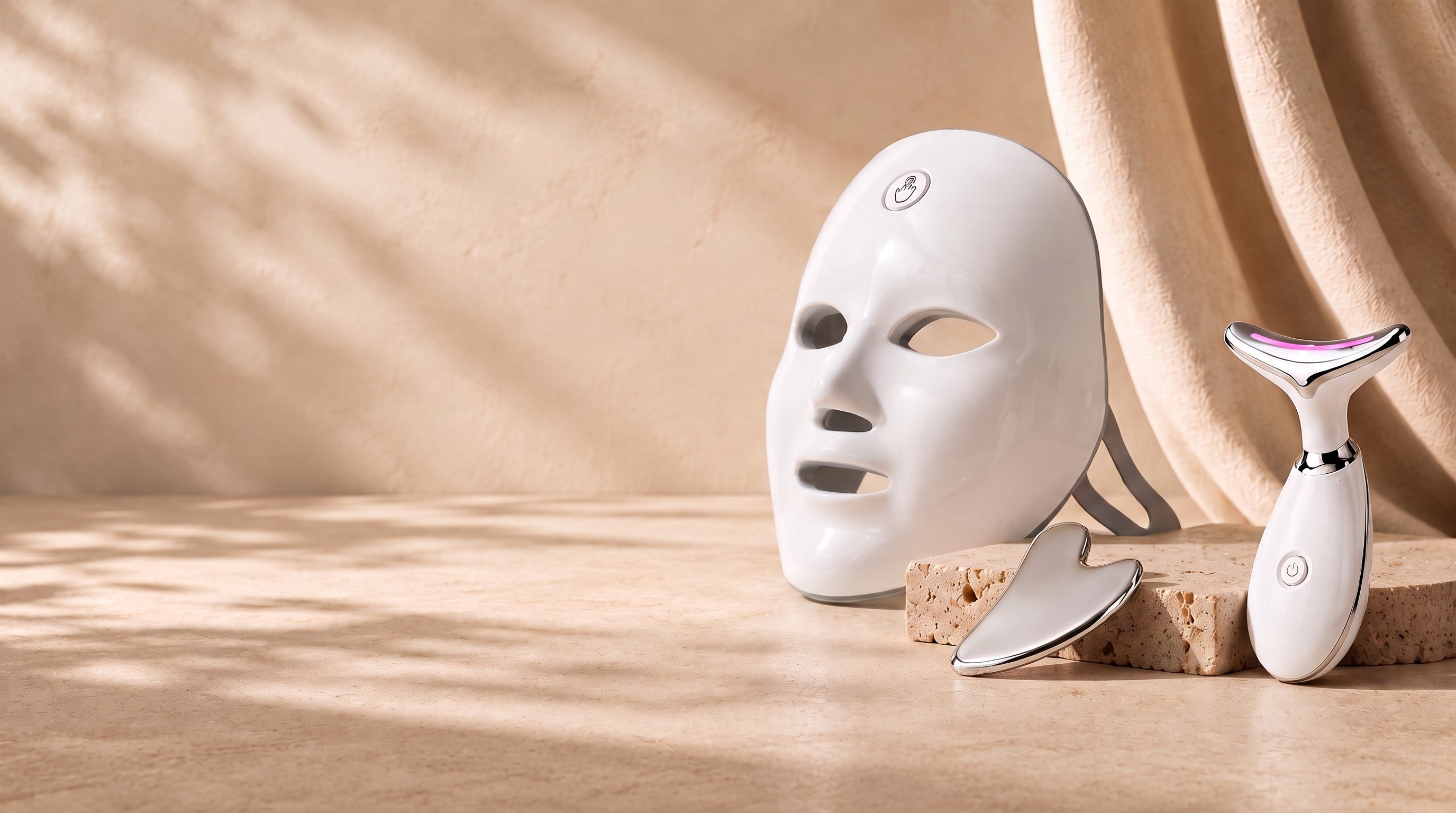 SENVÈ 7-color LED mask, stainless steel gua sha and neck & face device on a travertine plinth with beige linen backdrop